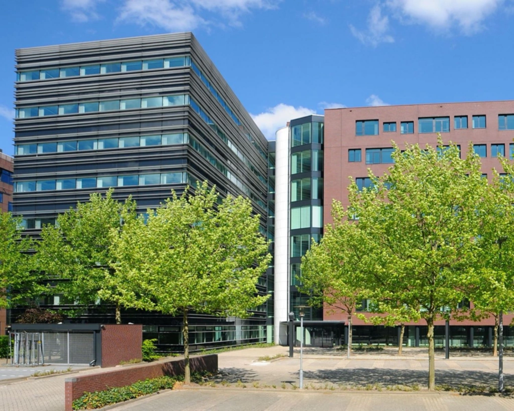 Modern kantoorgebouw met veel glas en baksteen, omgeven door jonge groene bomen en een lege parkeerplaats.