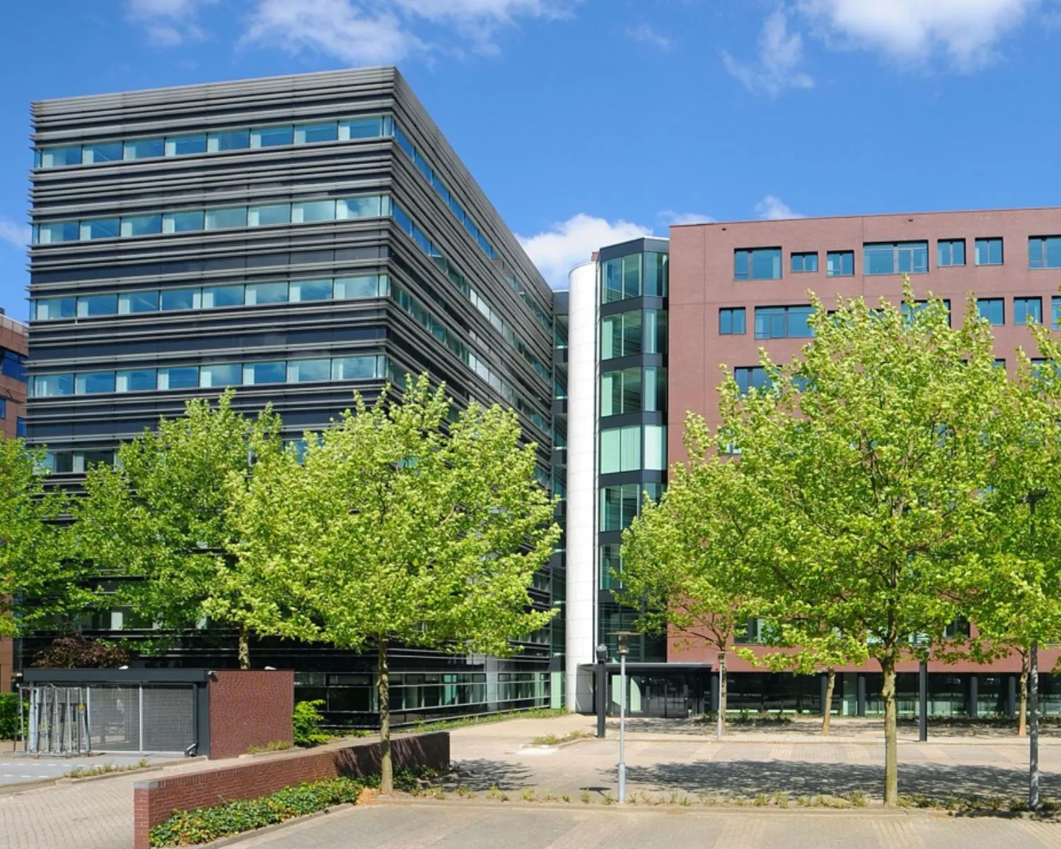 Modern kantoorgebouw met veel glas en baksteen, omgeven door jonge groene bomen en een lege parkeerplaats.