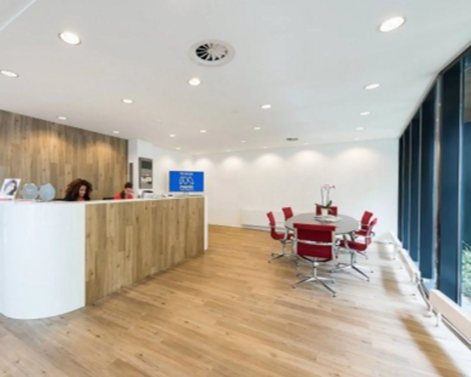 Moderne wachtruimte met houten vloer, ronde vergadertafel met rode stoelen en een receptiebalie met twee medewerkers.