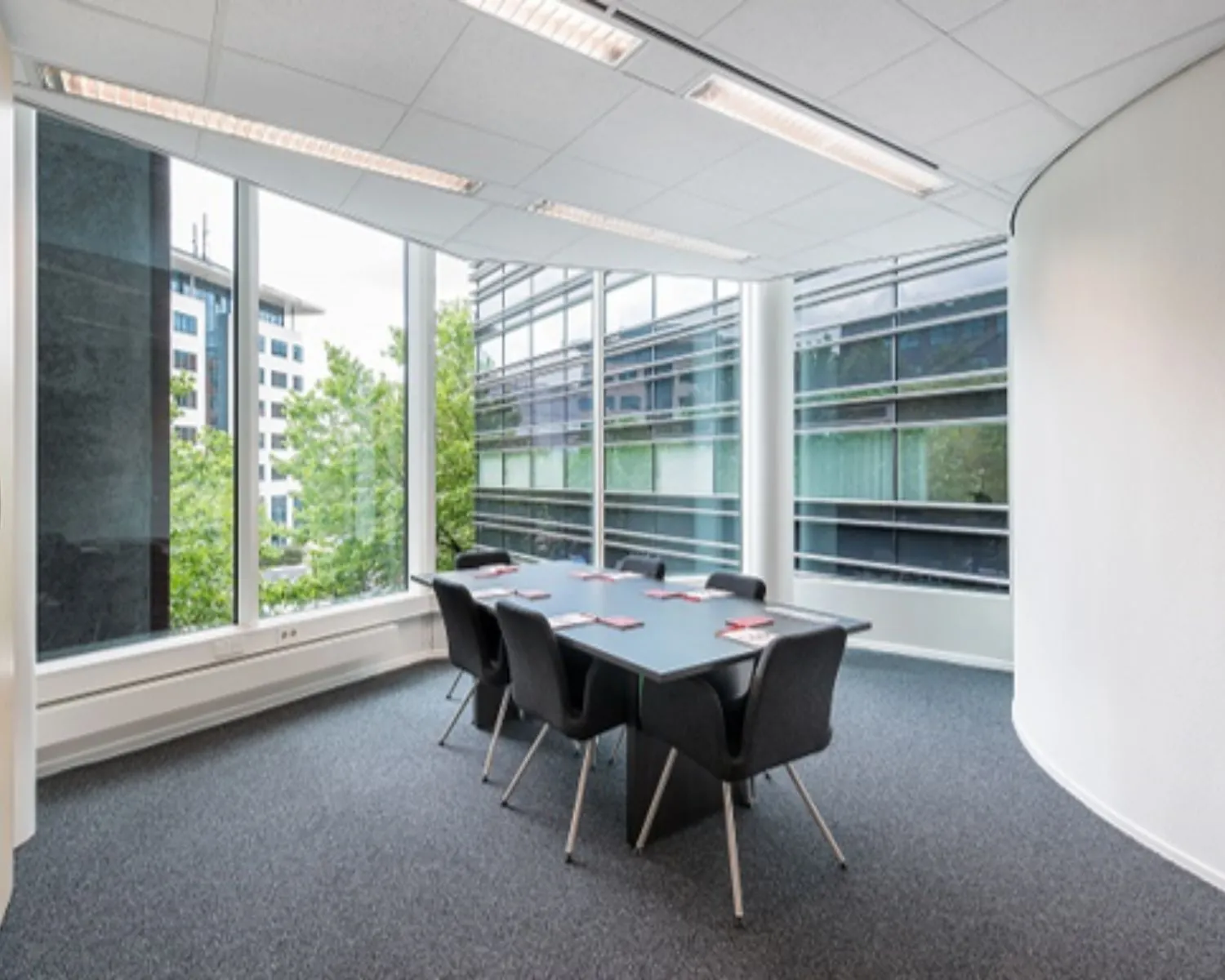 Moderne vergaderruimte met zes stoelen en grote ramen met uitzicht op kantoorgebouwen.