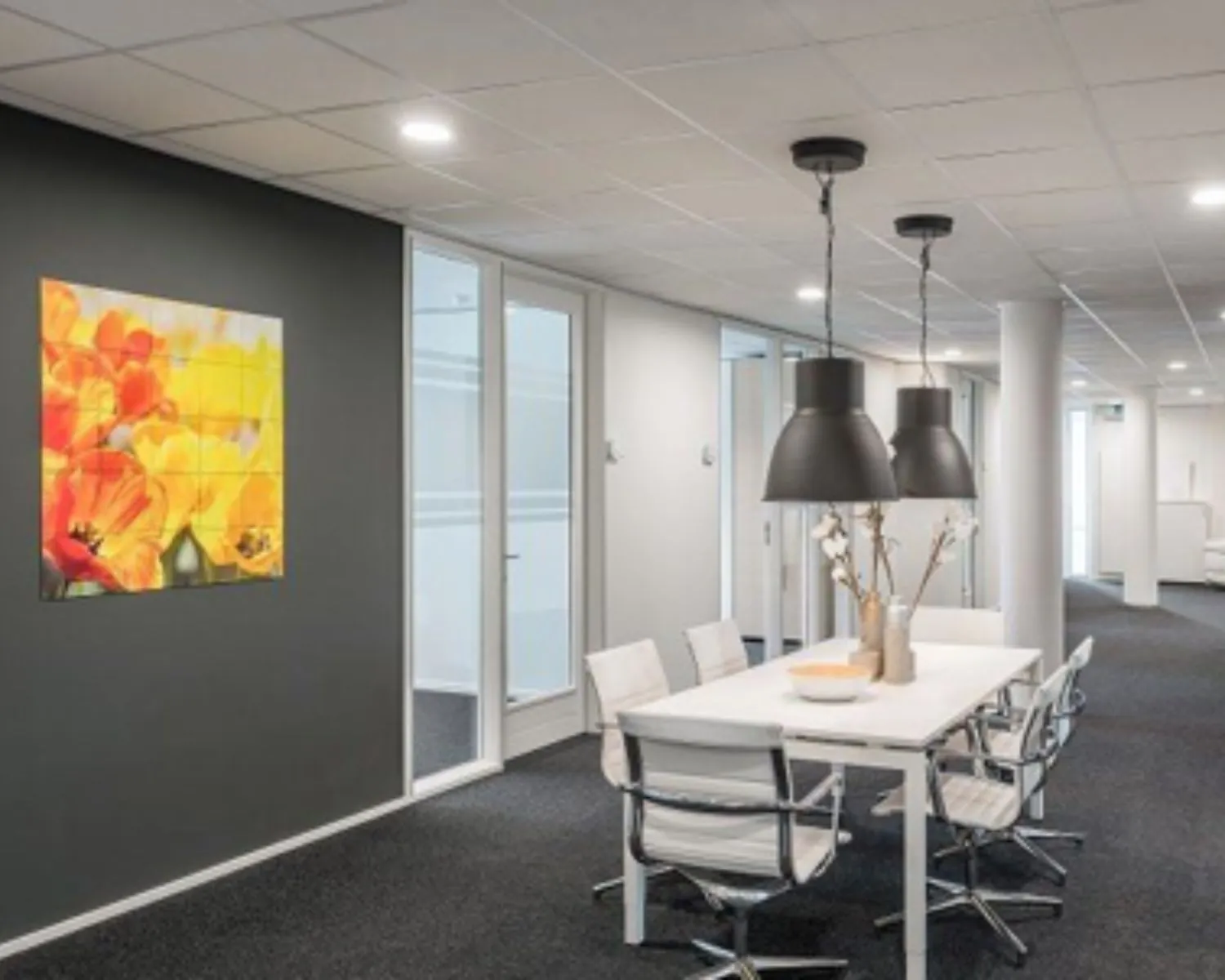 Moderne kantoorruimte met een witte vergadertafel, designstoelen, hanglampen en een kleurrijk bloemenkunstwerk aan de muur.