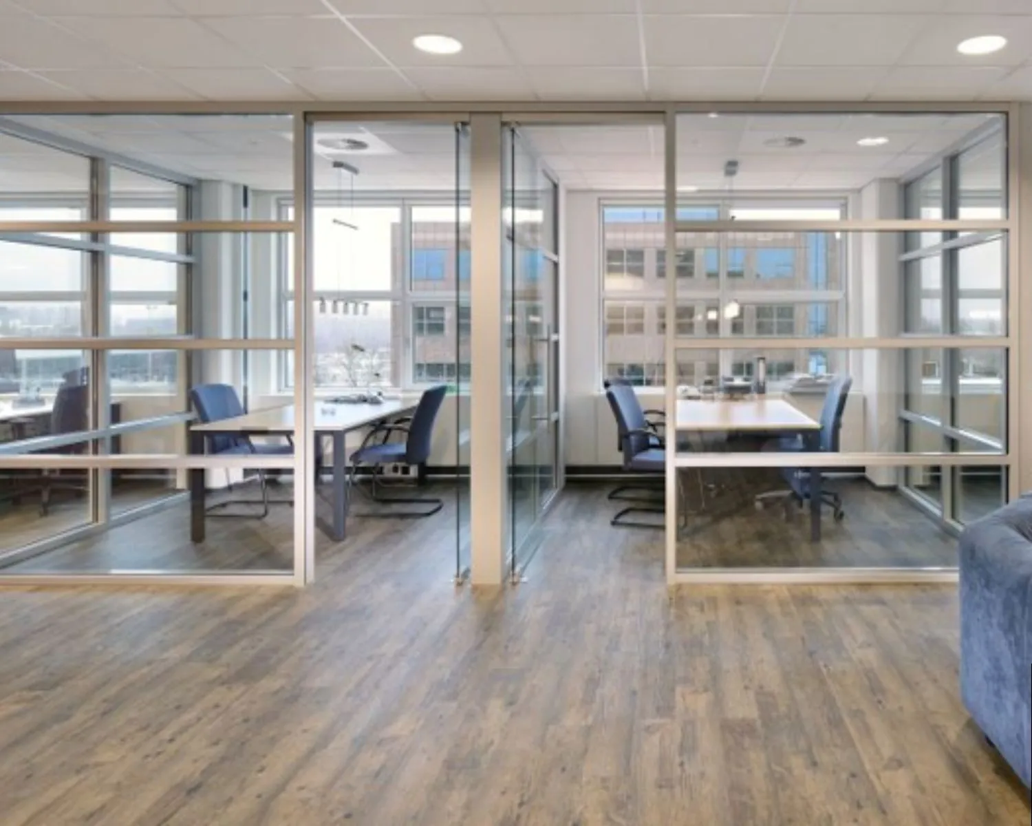Moderne kantoorruimte met glazen wanden, houten vloer en vergadertafels met blauwe stoelen.
