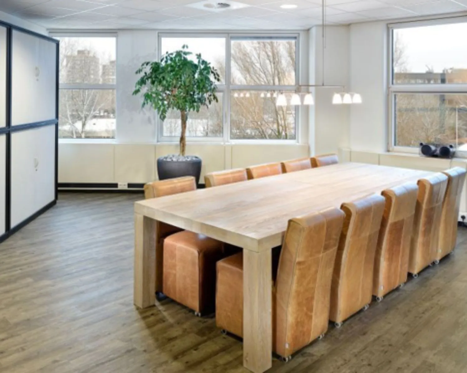 Vergaderruimte met een grote houten tafel, omringd door leren stoelen en natuurlijk licht vanuit meerdere ramen.