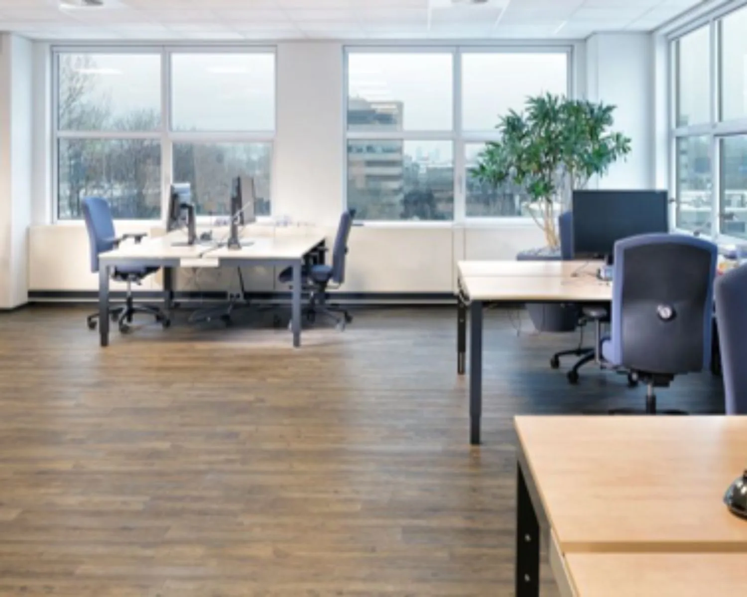 Ruim en modern kantoor met meerdere bureaus, bureaustoelen, computers en grote ramen met uitzicht op een stedelijk landschap.