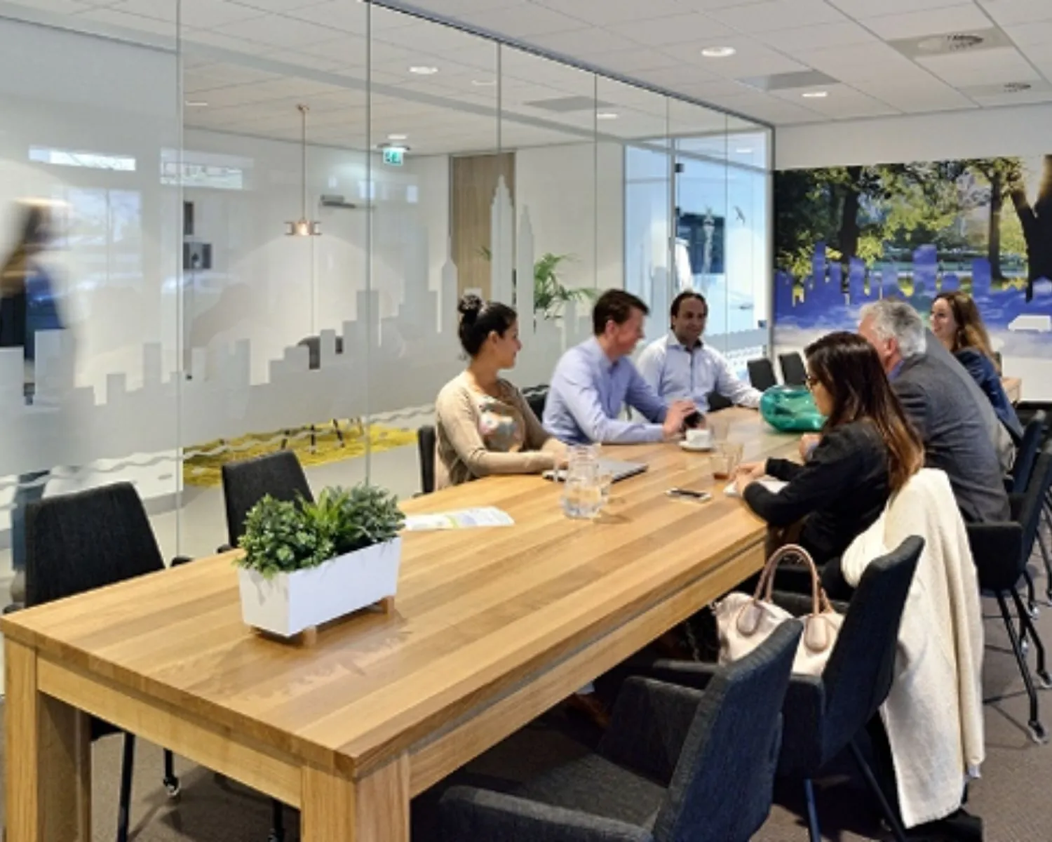 Zeven mensen zitten aan een houten vergadertafel in een moderne kantoorruimte met glazen wanden.