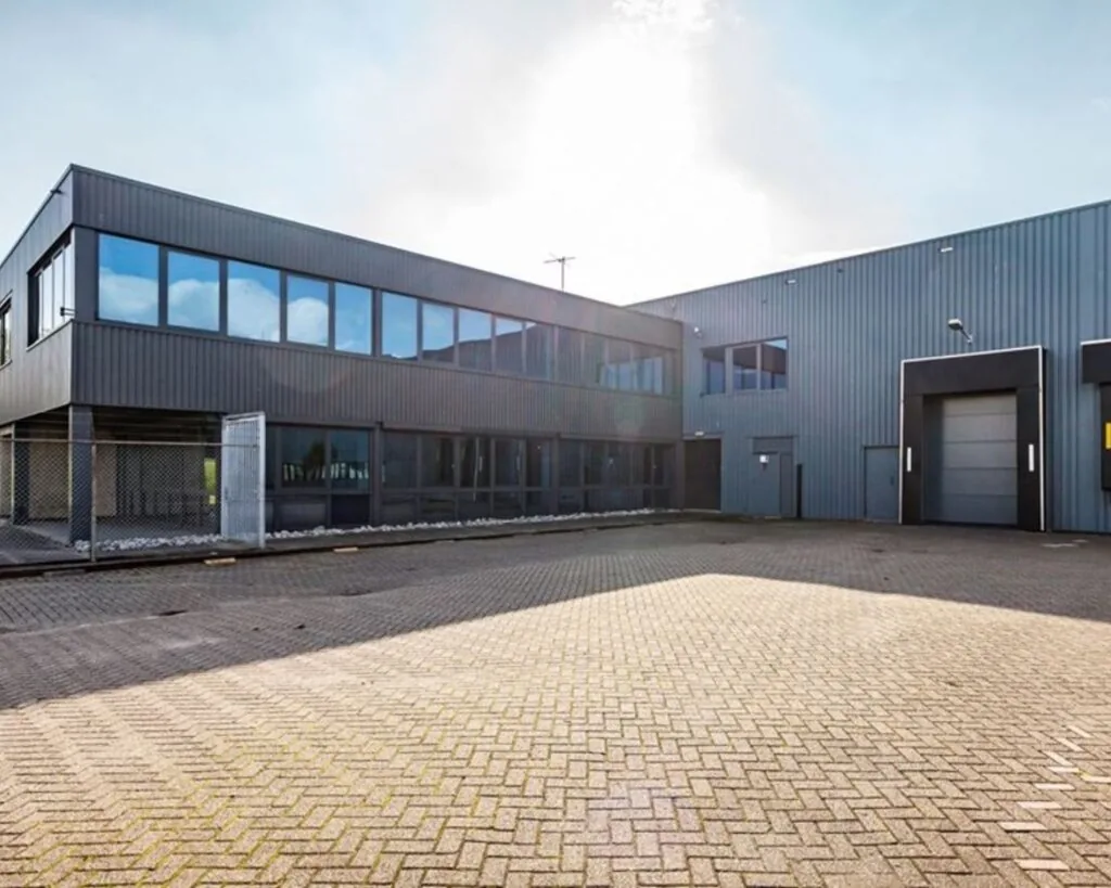 Grijze, moderne bedrijfsloods met grote ramen en rolluikdeur op een ruim, betegeld terrein.