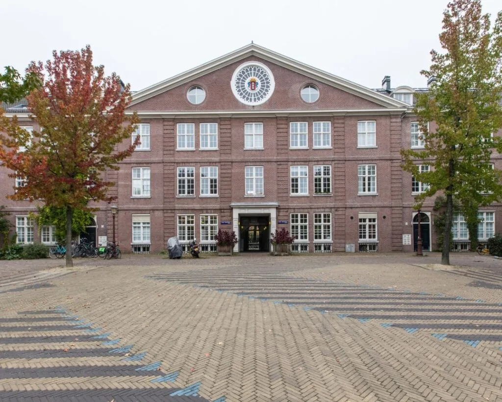 Vooraanzicht van een klassiek bakstenen schoolgebouw met symmetrische ramen en een ronde raampartij in de topgevel, geflankeerd door herfstkleurige bomen.