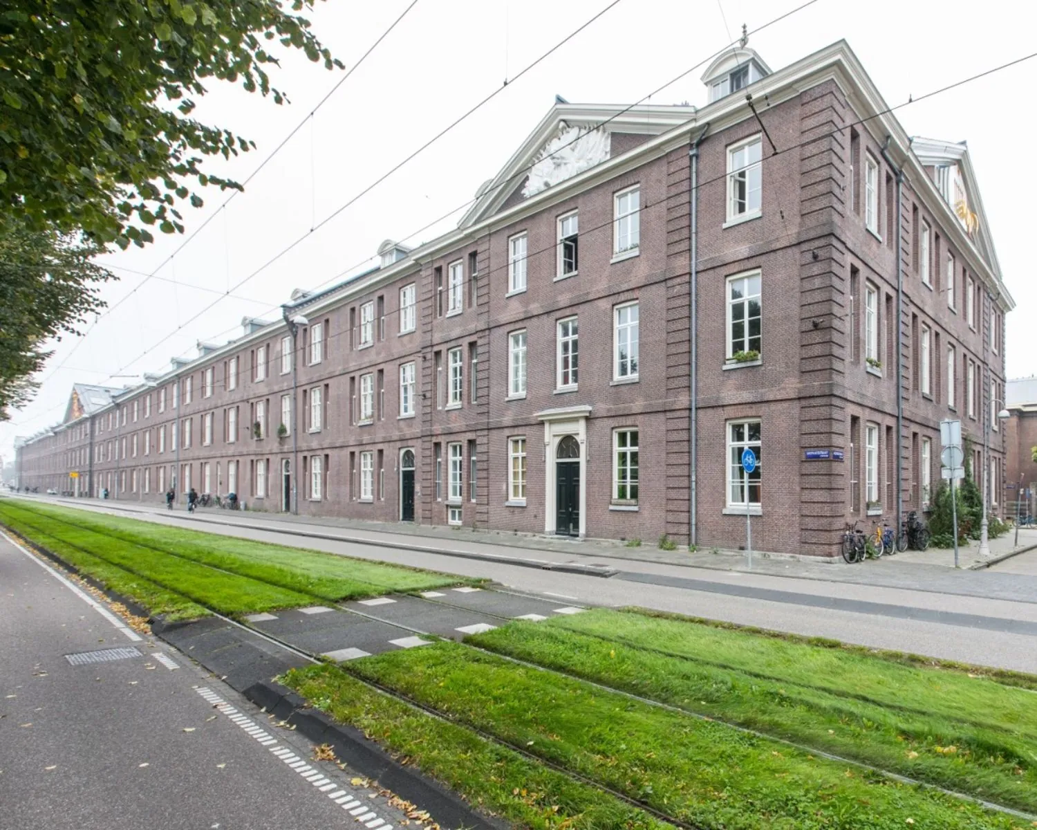 Langgerekt historisch bakstenen gebouw met witte kozijnen aan een straat met trambaan in een stedelijke omgeving.