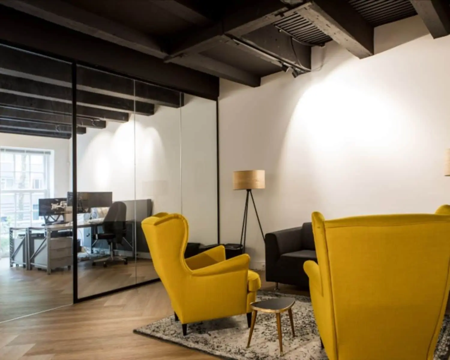 Moderne kantoorruimte met gele fauteuils, een zwarte bank en glazen wand die een werkruimte afscheidt.