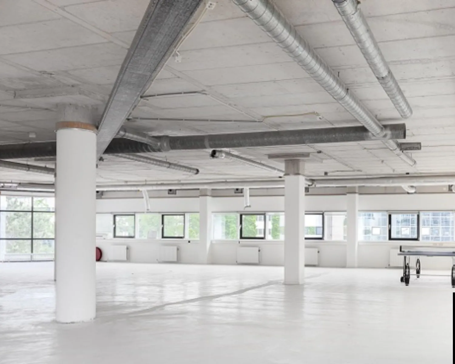 Licht en ruim leeg kantoorpand met zichtbare ventilatiebuizen en grote ramen.