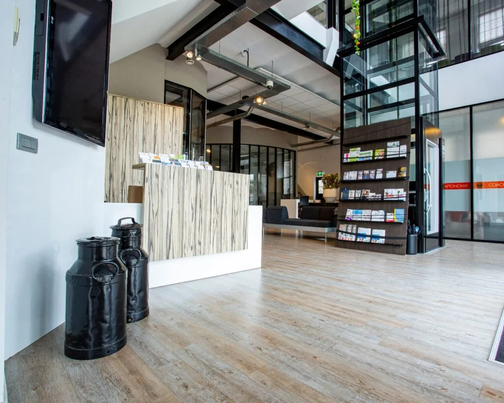 Moderne ontvangstruimte met een houten receptiebalie, folderrek, zitgedeelte en glazen lift.