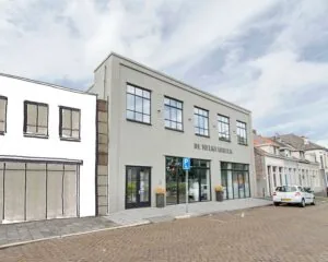 Vooraanzicht van het gebouw "De Melkfabriek" met grote ramen en een gehandicaptenparkeerplaats ervoor.