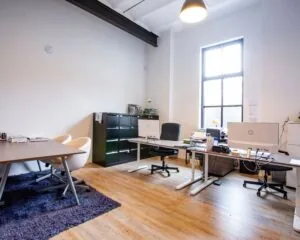 Modern ingerichte kantoorruimte met meerdere bureaus, computers en een groot raam dat natuurlijk licht binnenlaat.