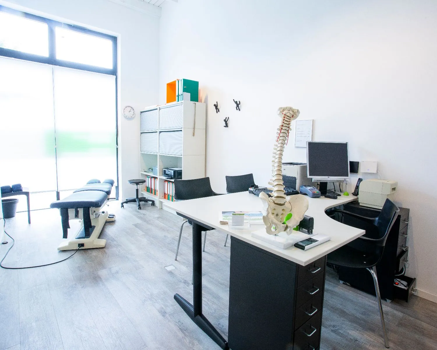 Moderne chiropractische behandelkamer met een behandelbank, bureau en wervelkolommodel.
