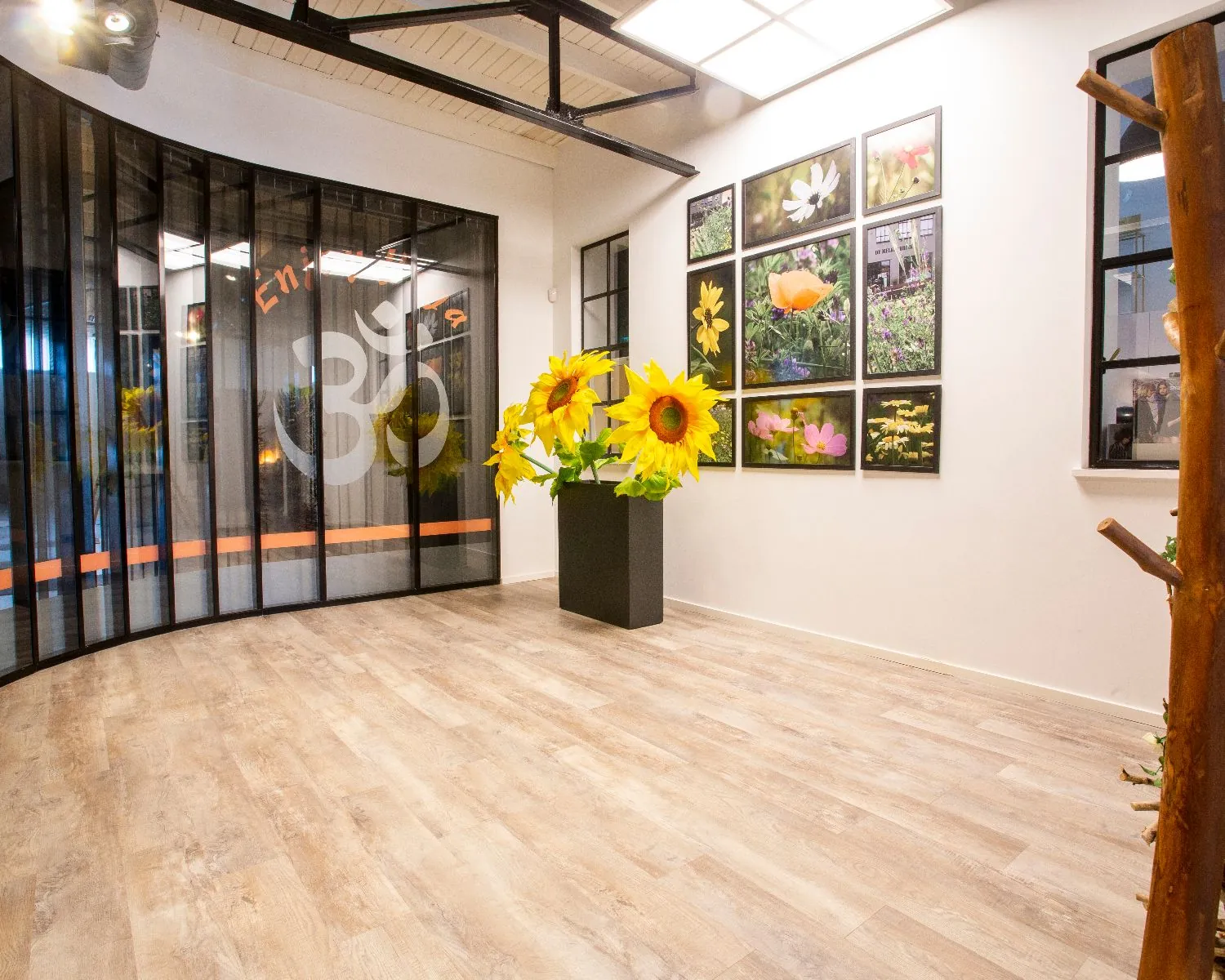 Heldere ontvangstruimte met houten vloer, een zwart gordijn met het Ohm-symbool, een vaas met grote zonnebloemen en een fotowand met kleurrijke bloemen.