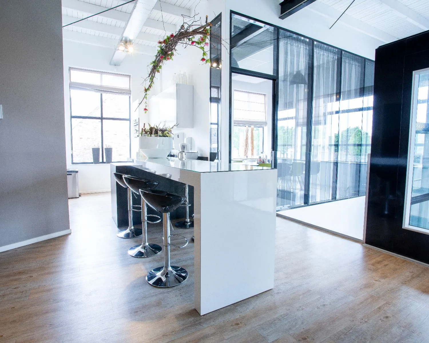 Moderne, lichte keukenruimte met witte wanden, zwarte barkrukken en glazen wanden.