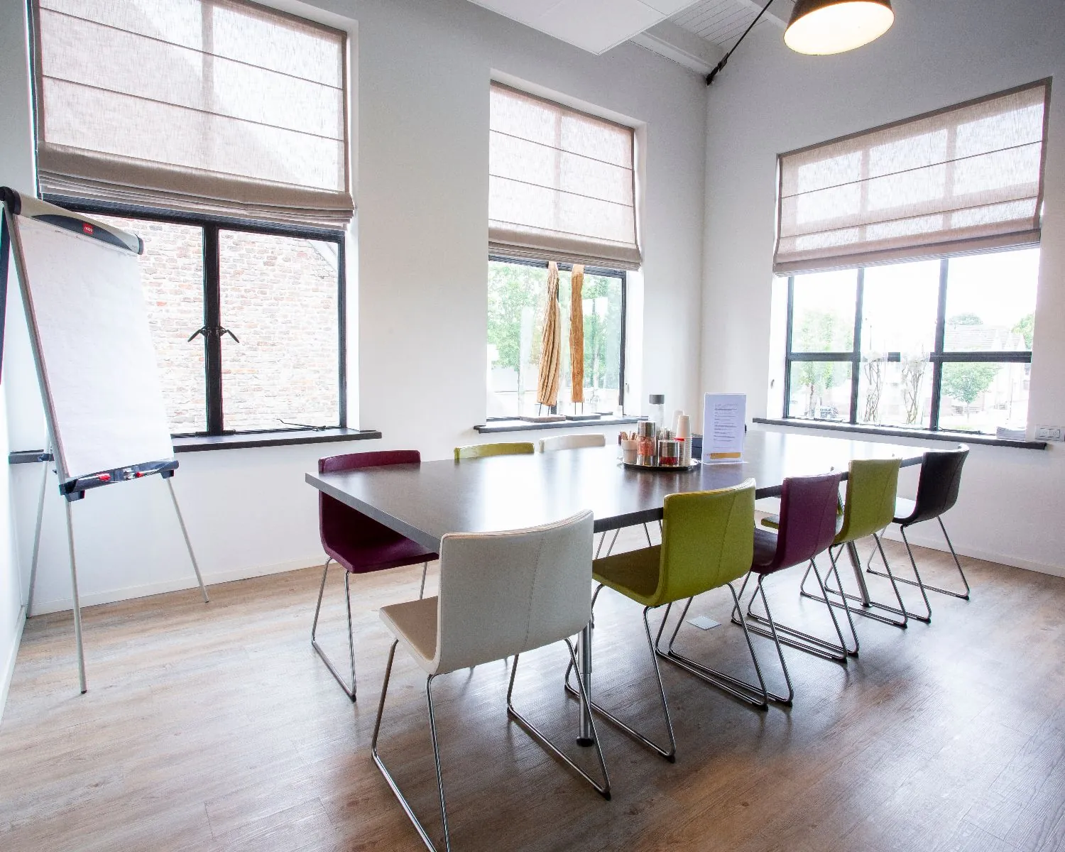 Moderne vergaderruimte met een lange tafel, kleurrijke stoelen, flip-over en grote ramen met veel daglicht.