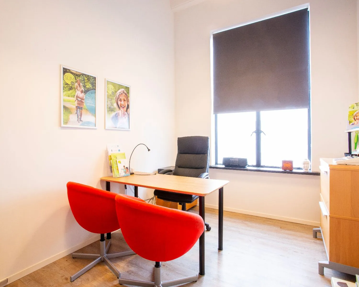 Moderne en lichte spreekkamer met houten bureau, twee rode stoelen en een zwart gordijn voor het raam.