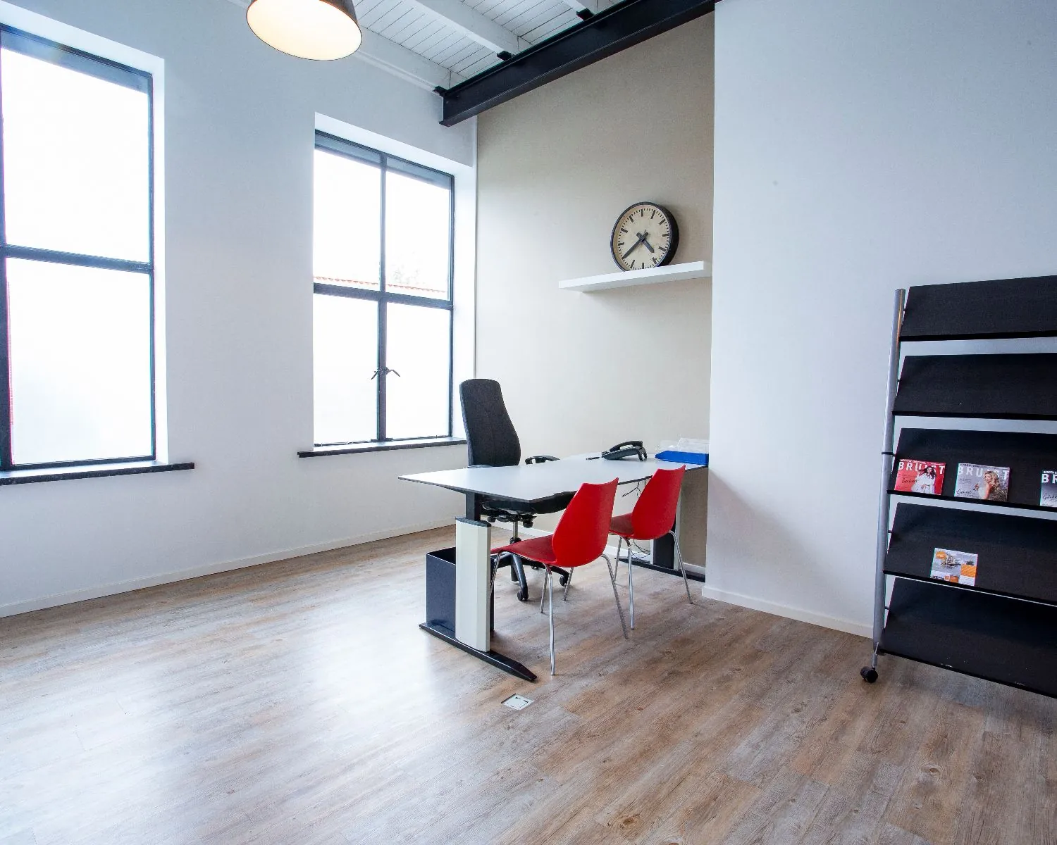 Modern ingericht kantoor met bureau, rode stoelen, tijdschriftrek en grote ramen.