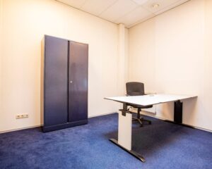 Kleine, eenvoudige kantoorruimte met een bureau, bureaustoel en een gesloten archiefkast.