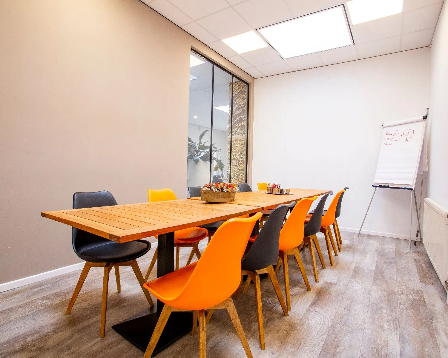Vergaderruimte met houten tafel, kleurrijke stoelen en een flip-over met notities.