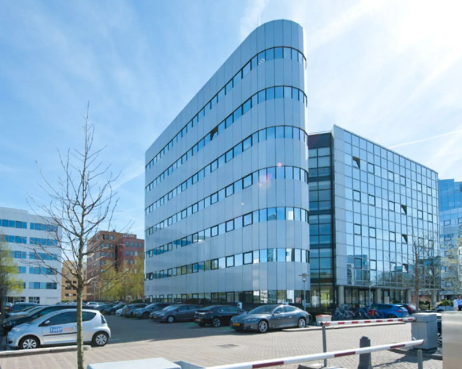 Modern kantoorgebouw met een ronde gevel en spiegelende ramen, met geparkeerde auto's op de voorgrond.