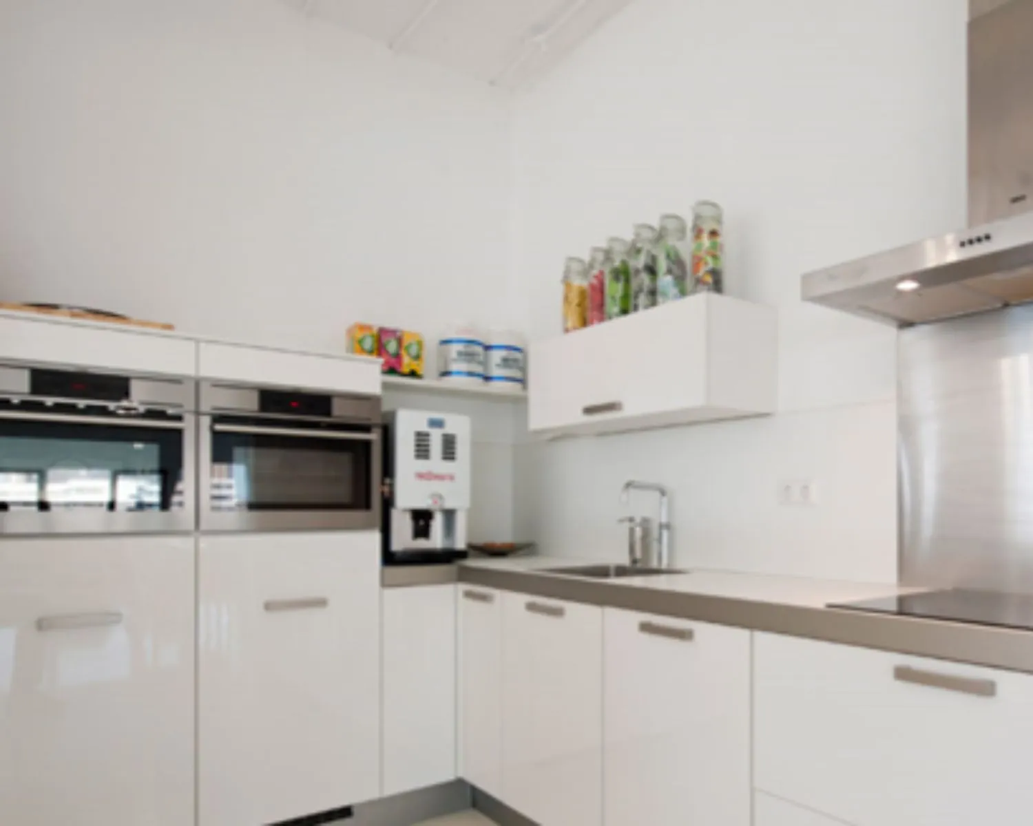 Moderne witte keuken met ingebouwde ovens, een koffiemachine en glazen potten gevuld met kleurrijke ingrediënten op een bovenkast.