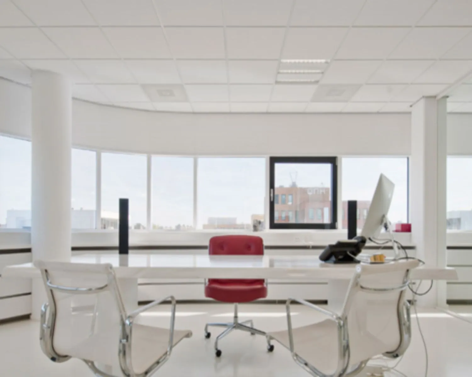 Modern en licht kantoor met grote ramen, een wit bureau, een rode bureaustoel en twee transparante stoelen.