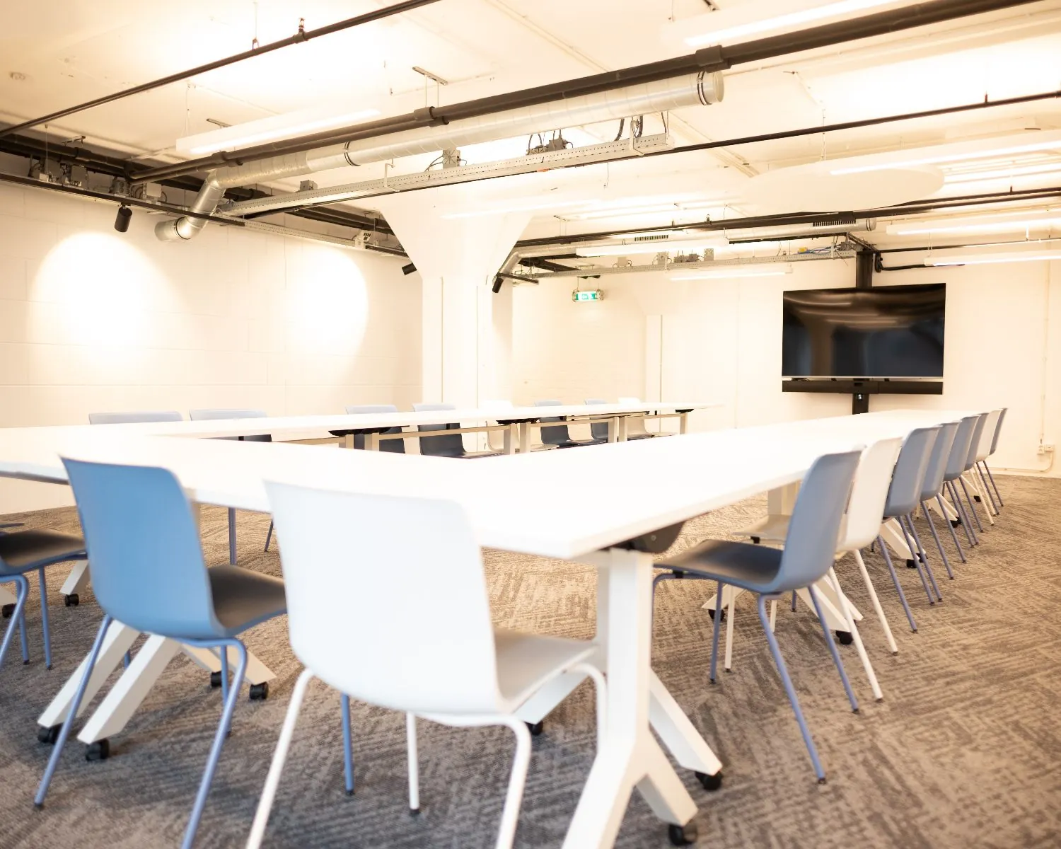 Moderne vergaderruimte met witte tafels, blauwe stoelen en een groot presentatiescherm aan de muur.