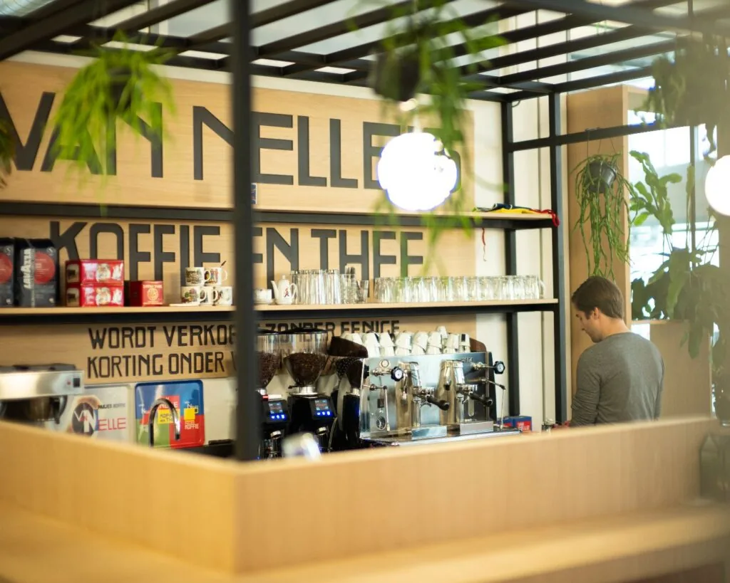 Barista bereidt koffie achter een moderne koffiebalie met Van Nelle-belettering op de achtergrond.