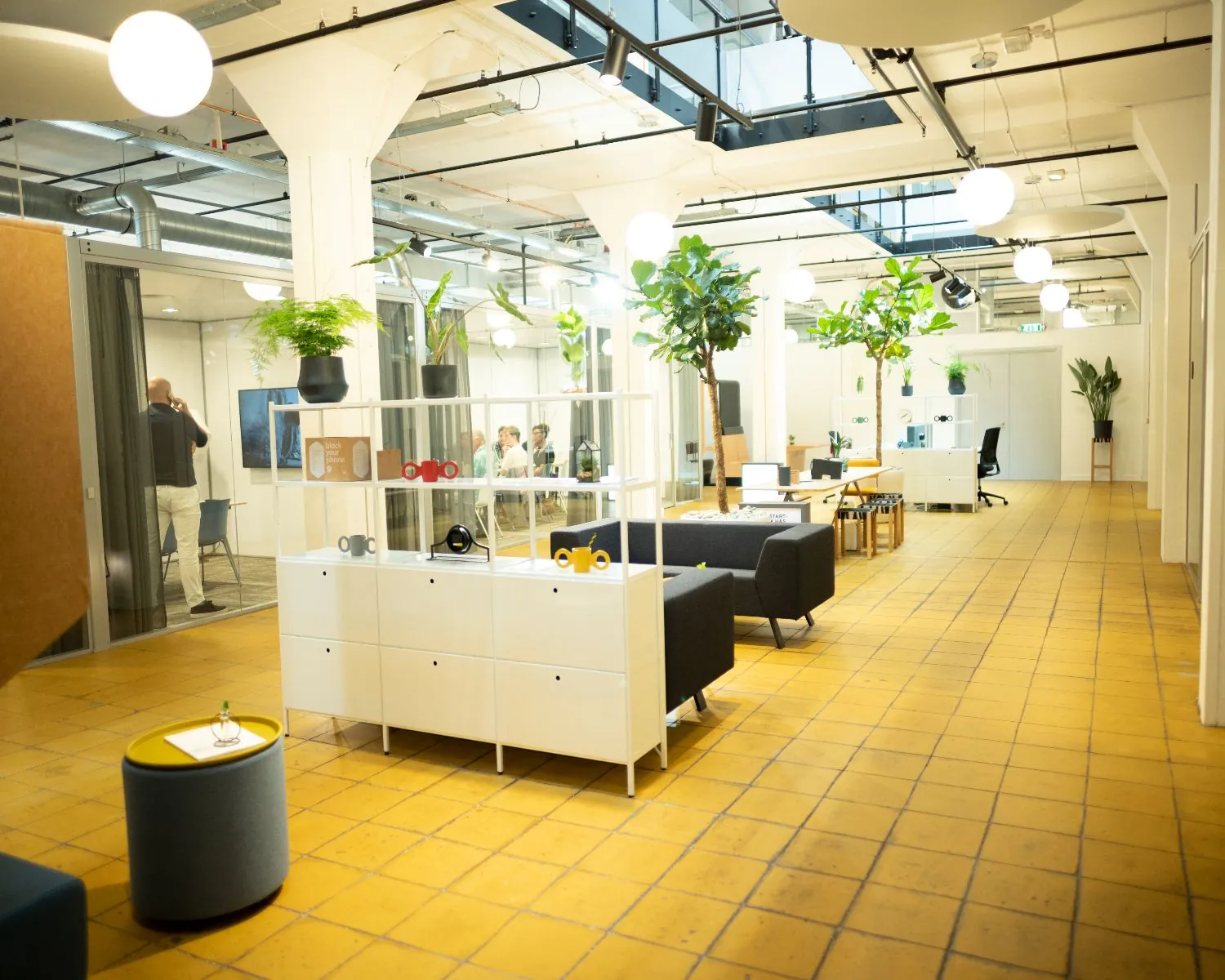 Modern ingerichte kantoortuin met glazen vergaderruimte, gele tegelvloer, planten en loungemeubilair.