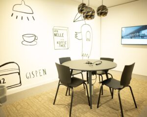 Vergaderruimte met ronde tafel, vier stoelen, wandtekeningen en een beeldscherm aan de muur.