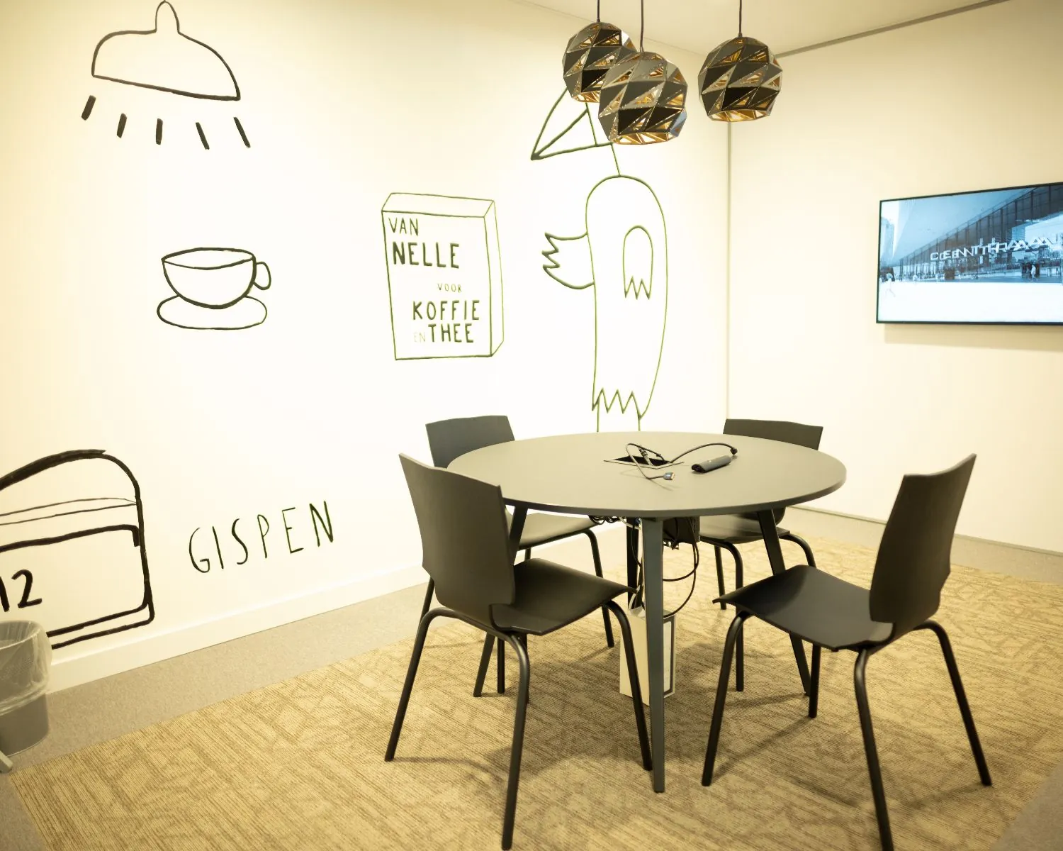 Vergaderruimte met ronde tafel, vier stoelen, wandtekeningen en een beeldscherm aan de muur.