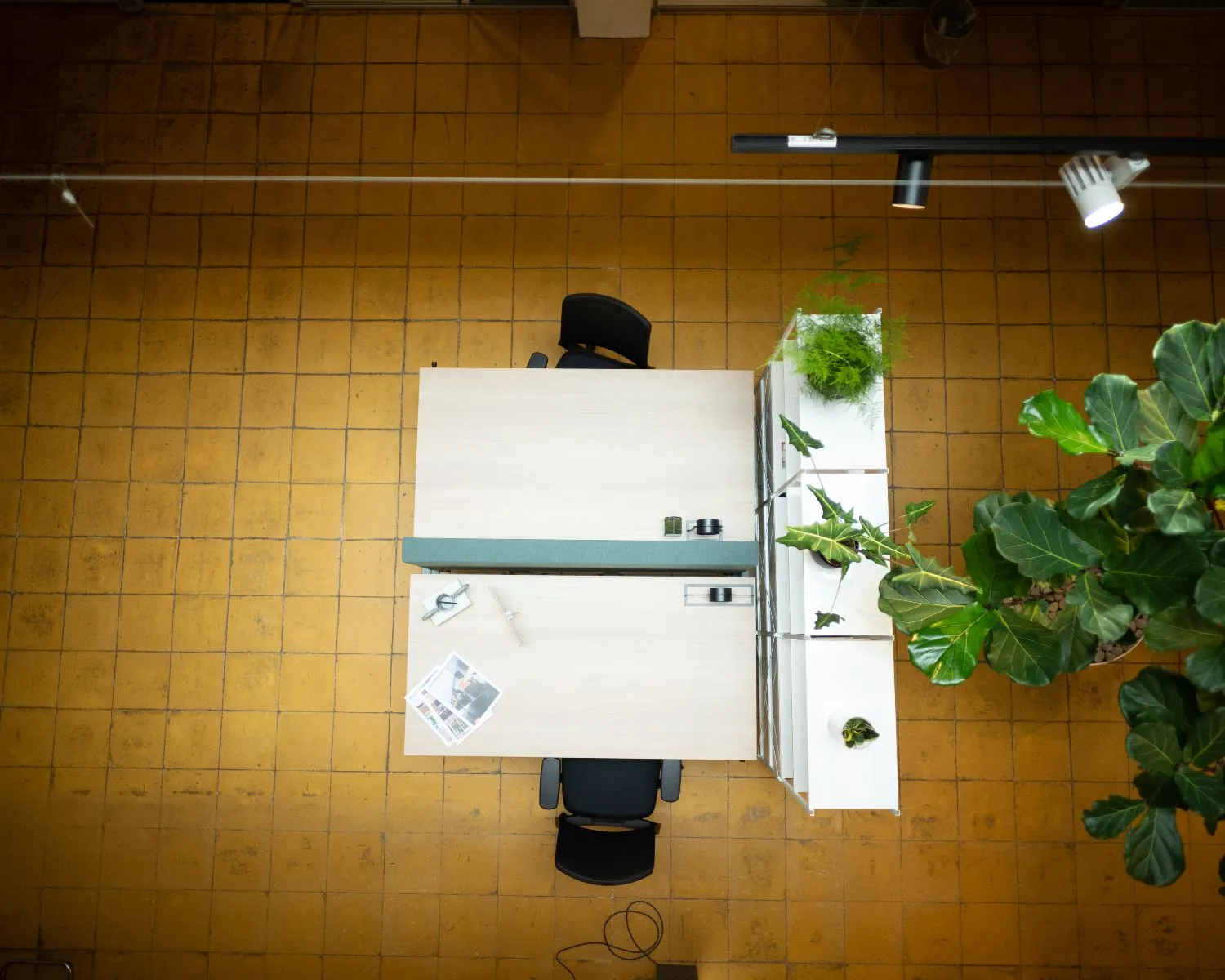 Bureaublad met twee werkplekken, omringd door planten, gezien van bovenaf op een vloer met gele tegels.