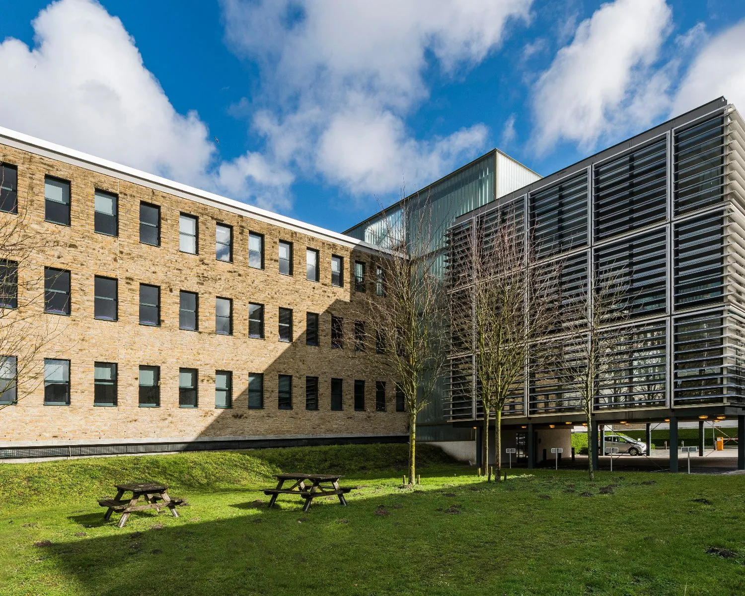 Modern kantoorgebouw met een bakstenen gevel en glazen aanbouw, met picknicktafels op het grasveld ervoor.