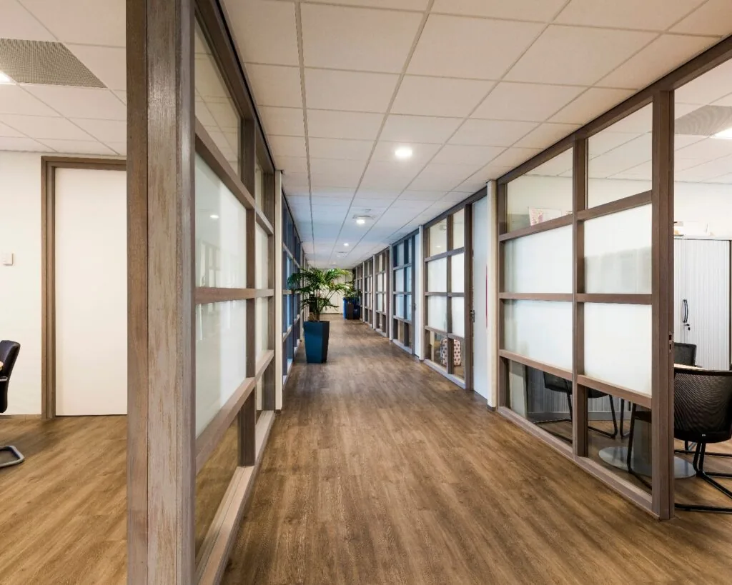 Moderne kantoorgang met glazen wanden, houten vloer en planten in blauwe potten.