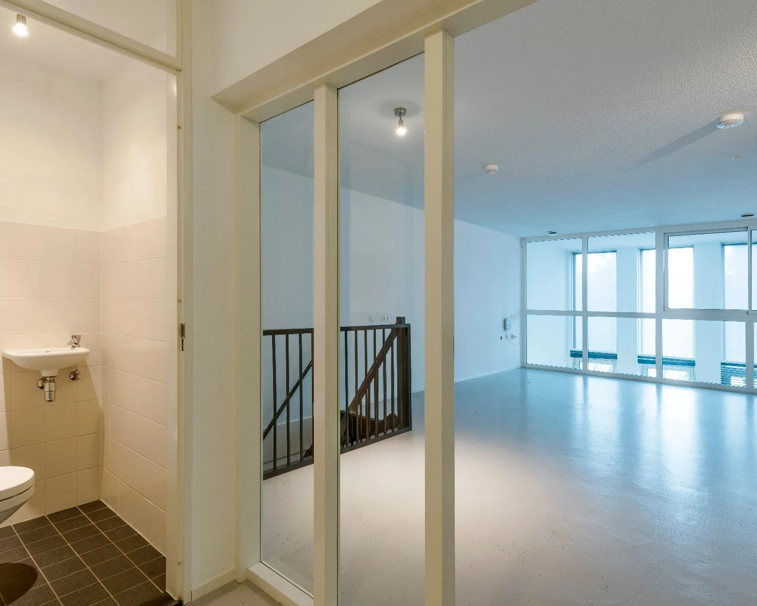 Lege, moderne binnenruimte met grote ramen, een trap en een kleine toiletruimte met wastafel.