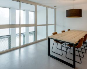 Vergaderruimte met een grote houten tafel, acht bruine stoelen en een zwarte hanglamp, omgeven door glazen wanden.