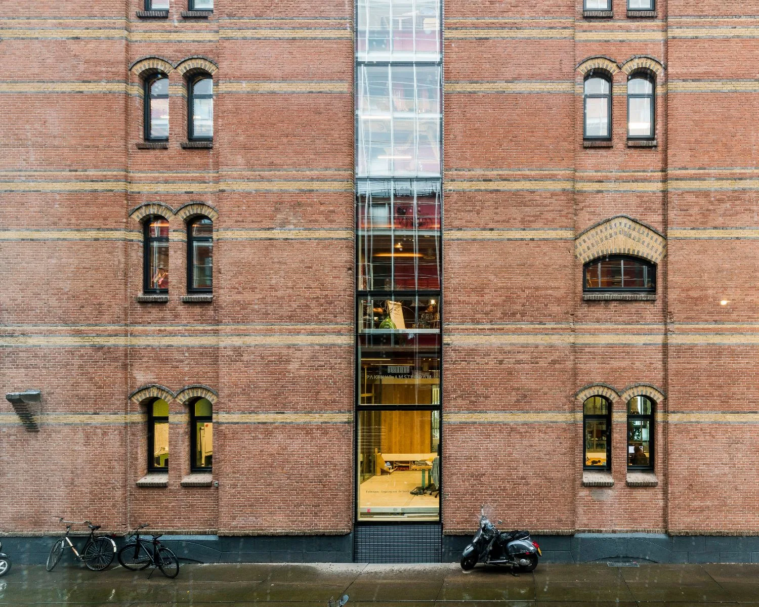 Bakstenen gevel van een historisch pand met een moderne glazen doorgang in het midden, fietsen en een scooter geparkeerd op de natte stoep.
