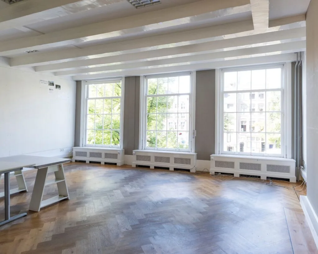 Lichte, lege kantoorruimte met houten visgraatvloer en grote ramen die uitkijken op een stadsomgeving.