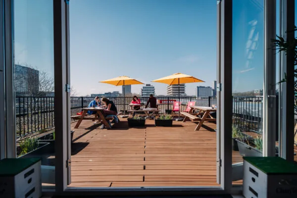 Zonnig dakterras bij KBF met picknicktafels, gele parasols en mensen die werken of ontspannen.
