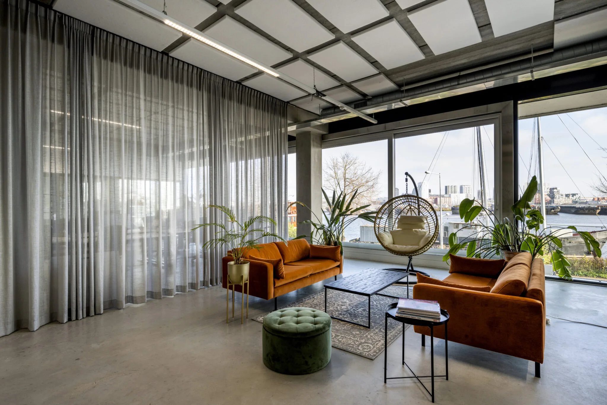 Moderne loungeruimte aan de Danzigerkade met oranje fluwelen banken, grote planten, een hangstoel en uitzicht op het water.