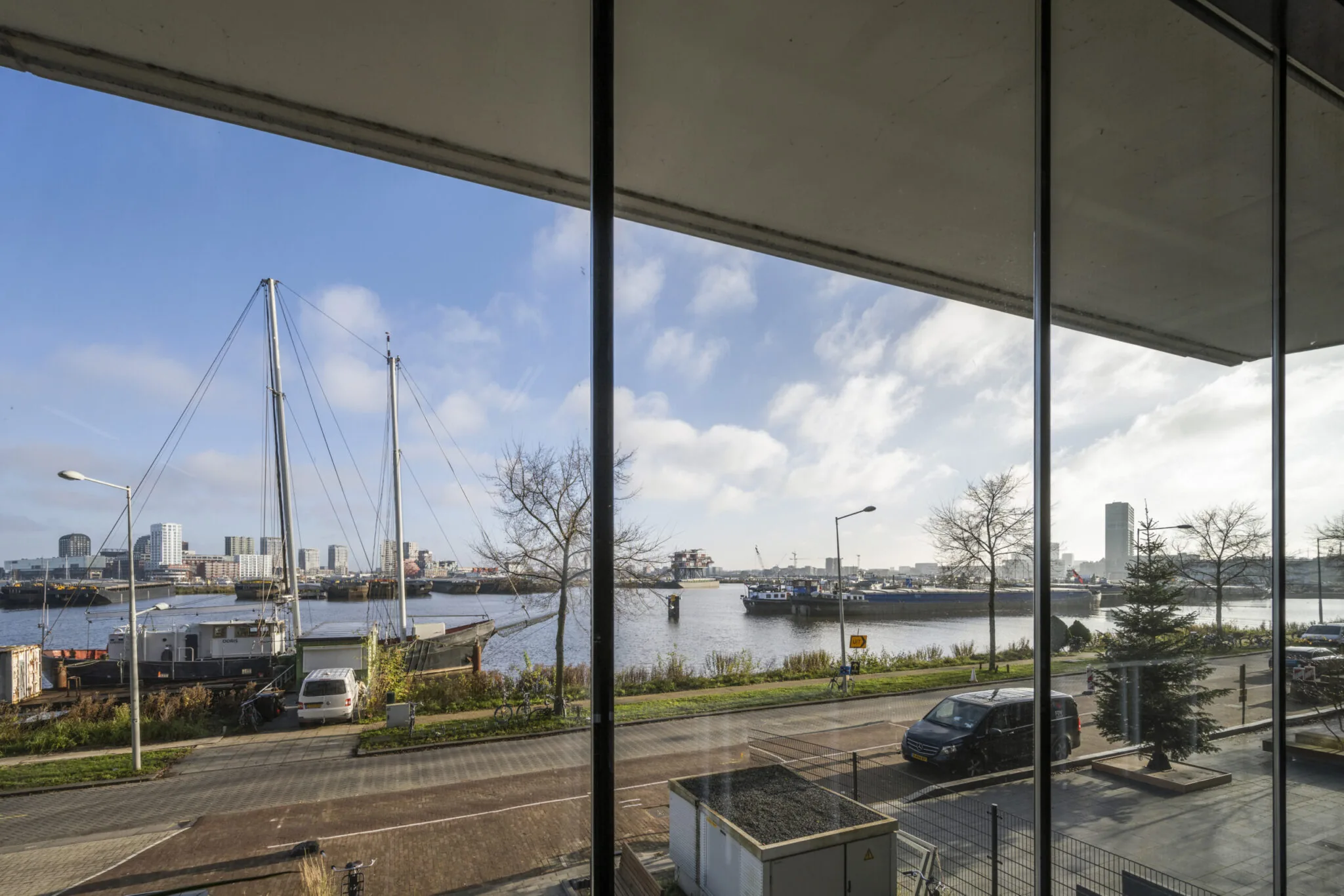 Uitzicht vanaf Danzigerkade op het IJ met schepen, kade en hoogbouw aan de overkant onder een blauwe lucht.