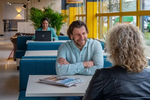 Twee mensen in gesprek aan een tafel in een moderne kantooromgeving aan de Disketteweg 14, met een derde persoon werkend op een laptop op de achtergrond.
