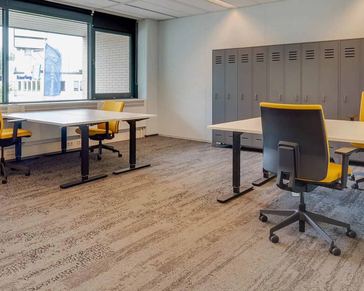 Modern kantoorinterieur met verstelbare bureaus, geel-zwarte bureaustoelen en een rij grijze lockerkasten.