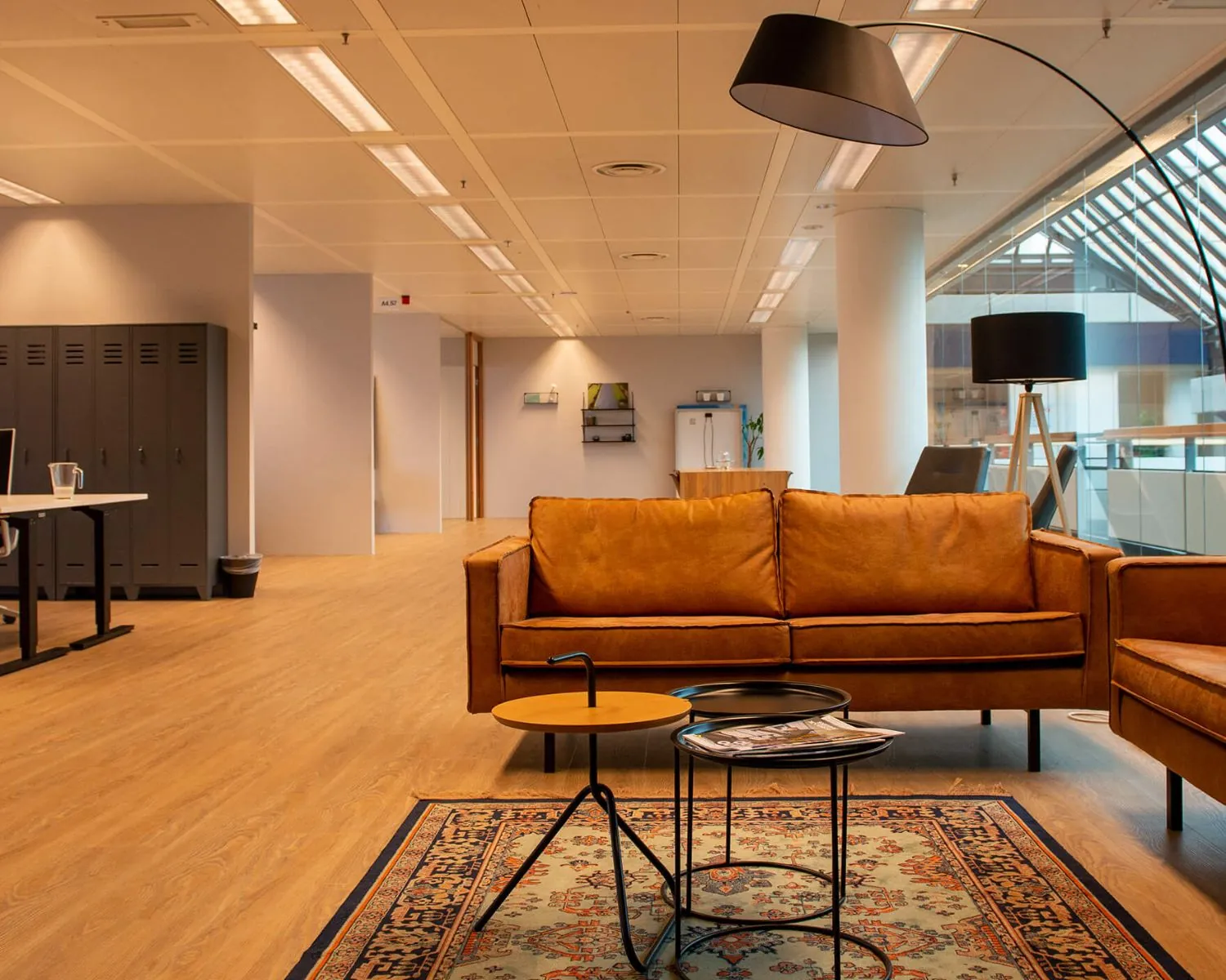 Modern kantoorinterieur met een bruine leren bank, vloerkleed, en designverlichting in een open werkruimte.