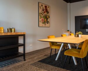 Moderne vergaderruimte met houten tafel, gele stoelen, floral schilderij en minimalistische decoratie.