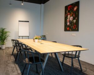 Vergaderruimte met houten tafel, zwarte stoelen, een flip-over en een schilderij met bloemen aan de muur.
