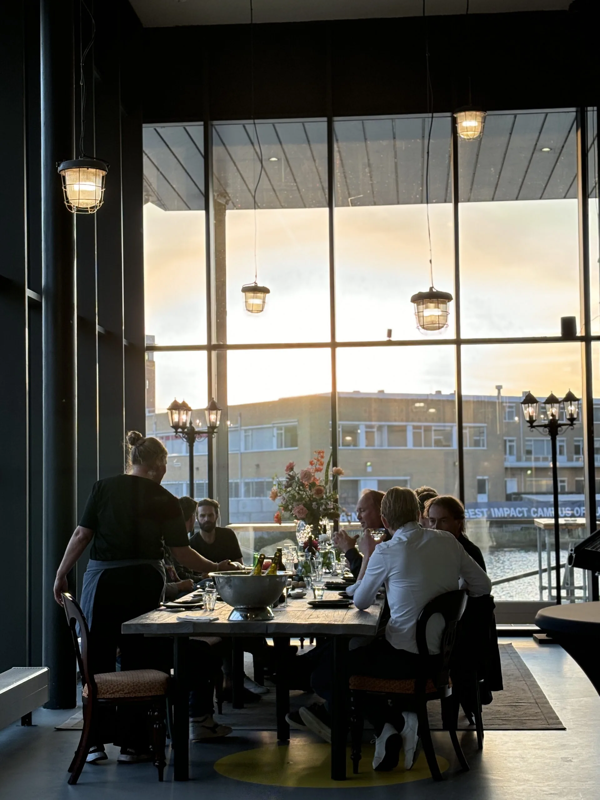 Een groep mensen dineert samen aan een sfeervol verlichte tafel bij zonsondergang in een moderne, glazen ruimte genaamd Focus.