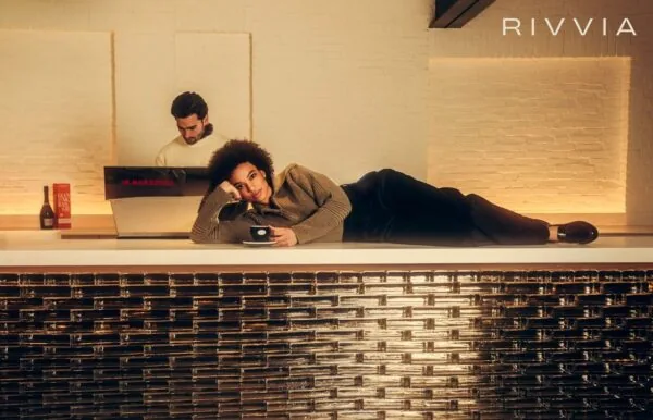 Vrouw ligt met een kopje koffie op een moderne bar terwijl een barista werkt achter een La Marzocco koffiemachine in een stijlvolle ruimte met het logo van RIVVIA op de muur.